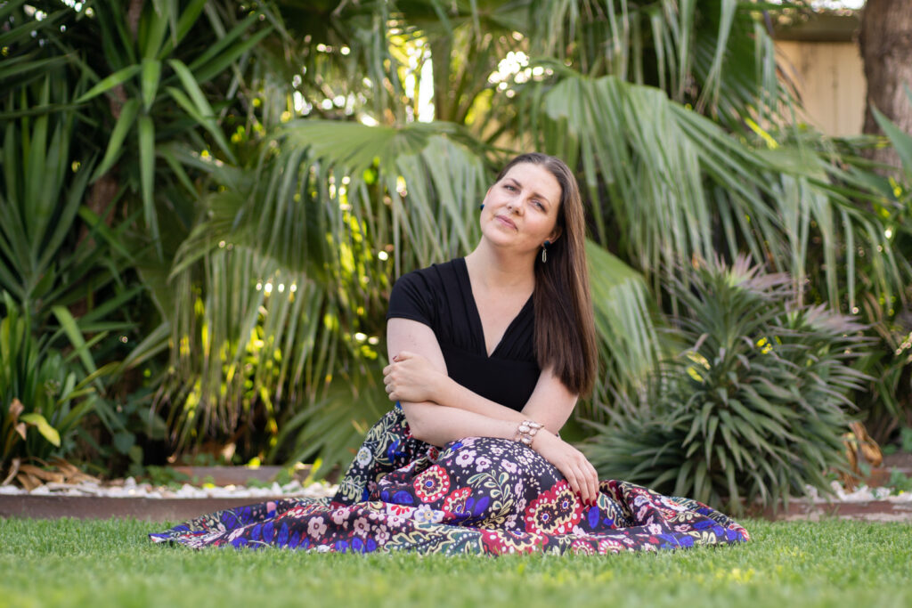 Australian travel writer Jessica Korteman sitting on the grass in front of a palm in Melbourne, Australia. She is wearing a blue patterned skirt and a black short-sleeved top. She is facing the camera, with her head tilted slightly to one side with long, brown hair.